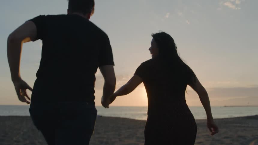 A man and woman hold hands while running along a beach together in the evening