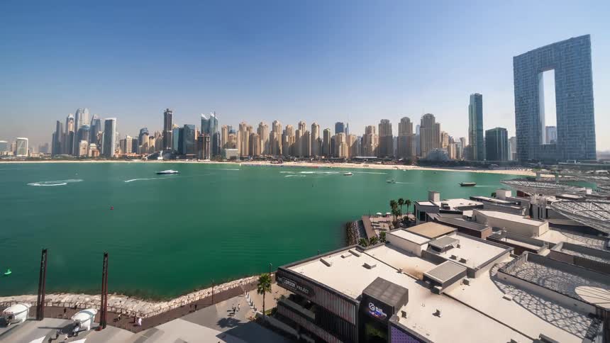 People move along coast and boats through water with city skyline in the background