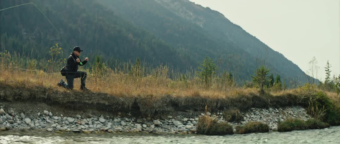 A man is kneeling at the side of a river as he throws his fishing line into the water
