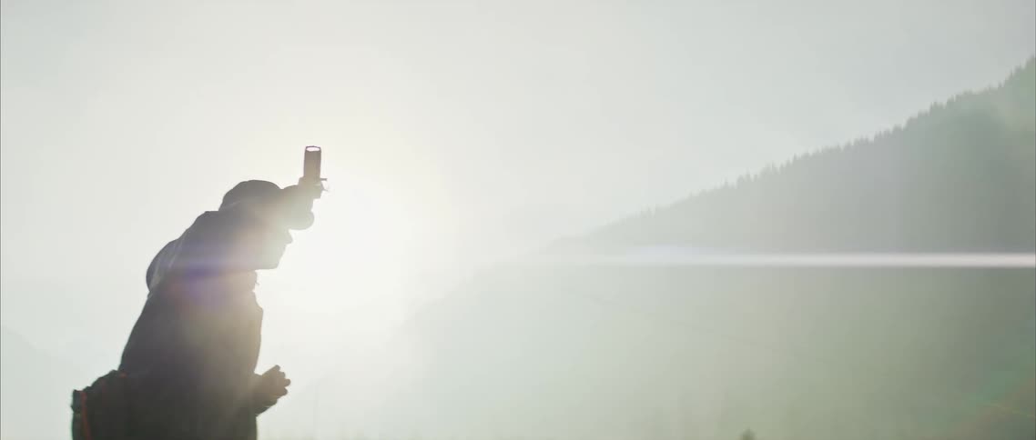 Man with black cap coat and pants casts fishing rod by river at sunrise