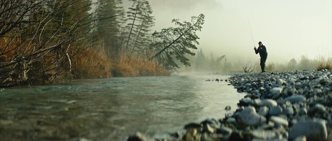 A man is fishing on a riverbank near a lot of trees