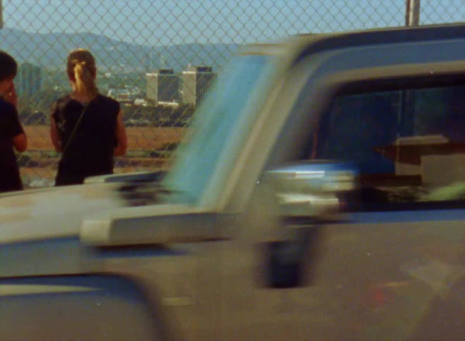 Trucks pass by people standing at fence watching grass fire with city and mountains in the background