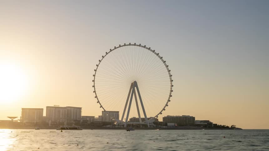A feris wheel is turning along the coast as boats are sailing in the water and city buildings are in the distance