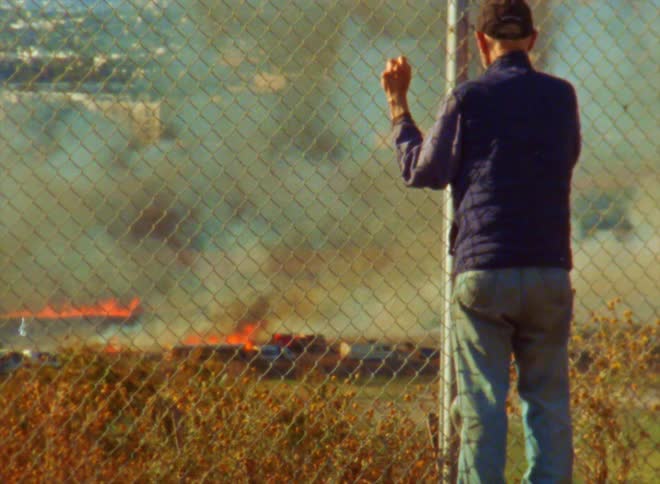 An elderly man stands against a fence and watches a fire burn in a field