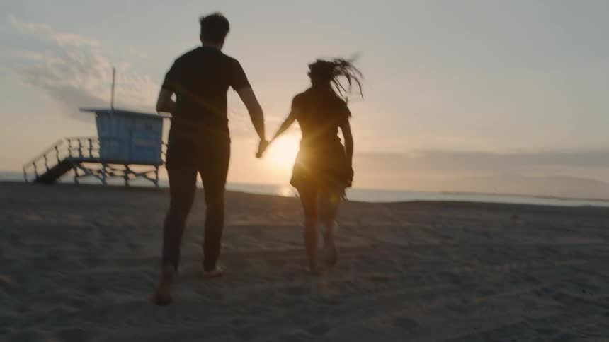 Couple runs across sandy beach toward sun sitting over water in a red sky
