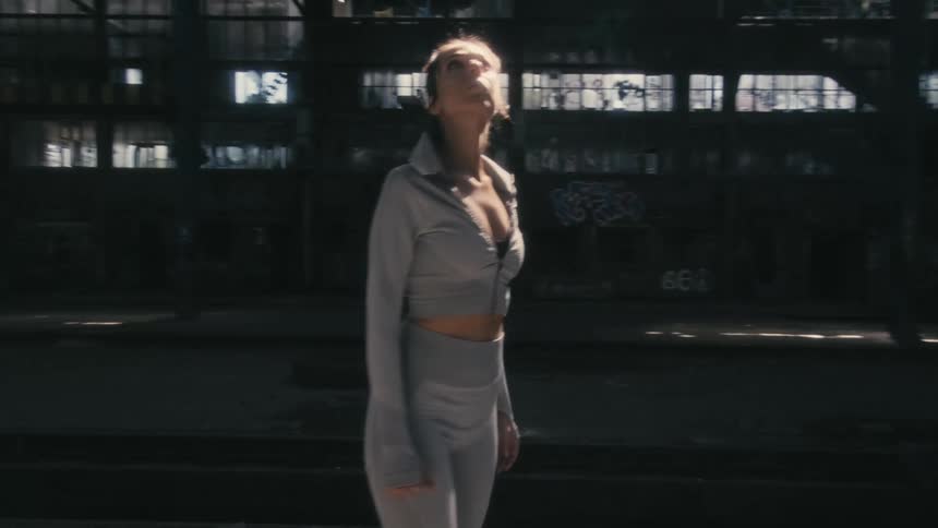 A woman is standing inside and abandoned building looking around as sunlight shines in from the windows