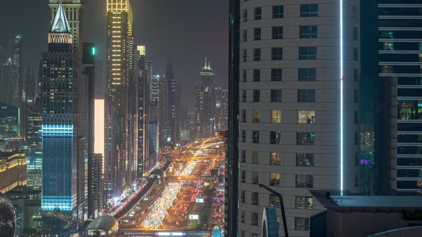 Dense traffic travels along a freeway located between skyscrapers in a metropolis at night