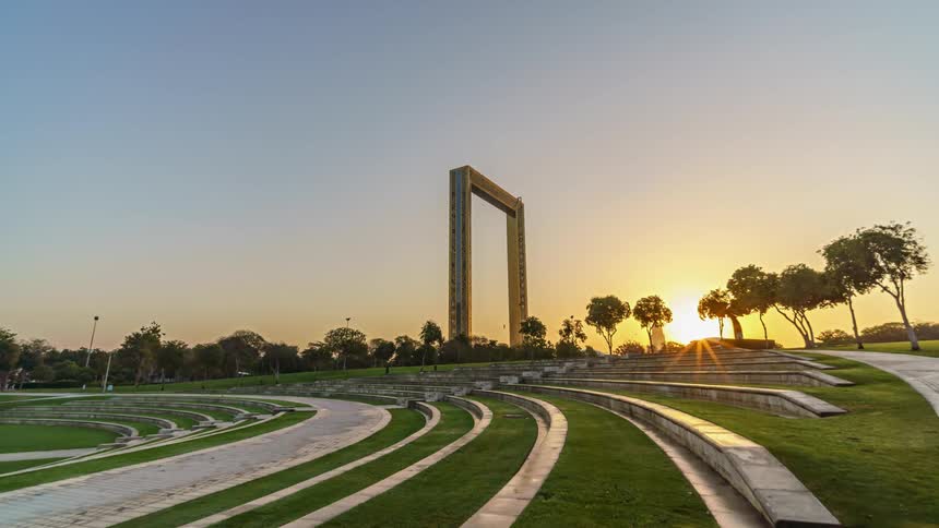 Walkways lead around the area ofthe dubai frame as it stands tall and the sun rises behind the trees in a clear sky