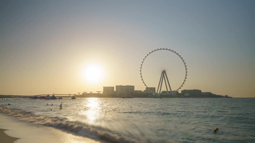People are walking on the beach and going into the water as a feris wheel and city skyline are in the distance and the sun is shining brightly in the sky
