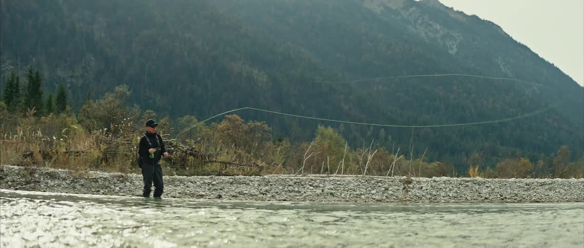 A man is standing at the side of a river as he casts his fishing line into the water