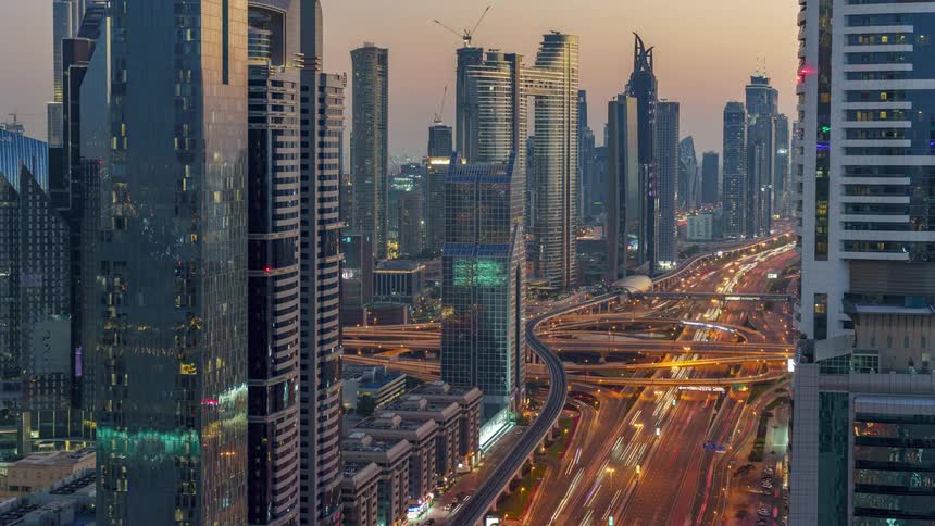 Traffic moves on the winding freeway below tall towers and futuristic buildings in the city of dubai as day becomes night and the city lights up