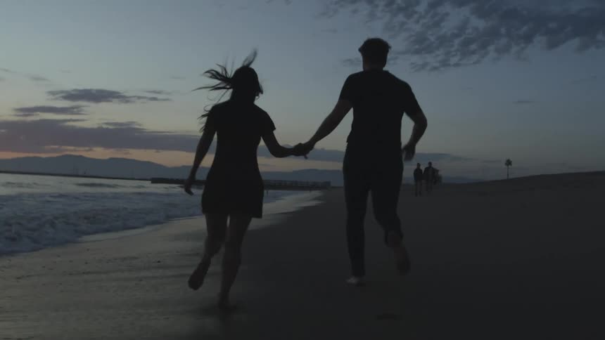 The silhouette of a couple holding hands and running on the beach at sunset
