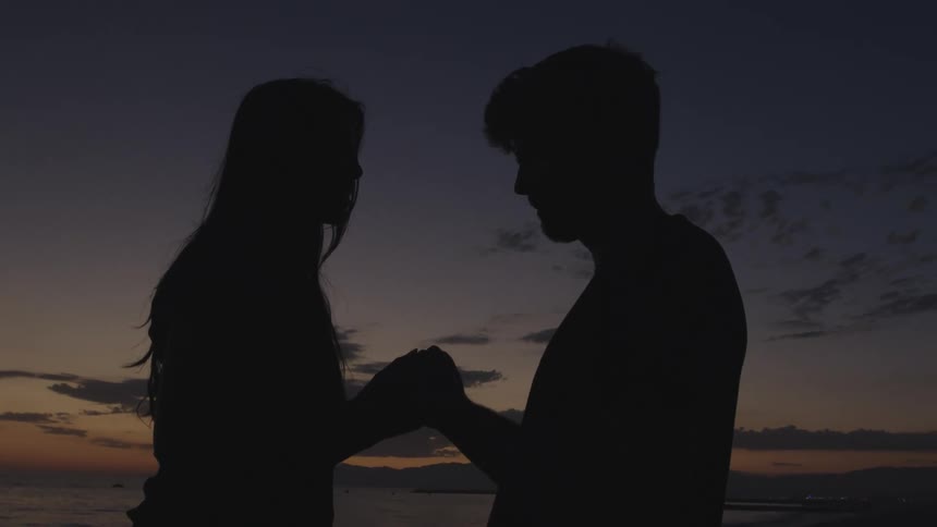Silhouette of a young couple holding hands while looking at each other at sunset next to the ocean
