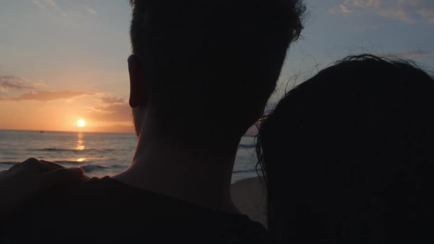 The silhouette of a woman leaning her head on her boyfriends shoulder as they watch the sun set over the ocean