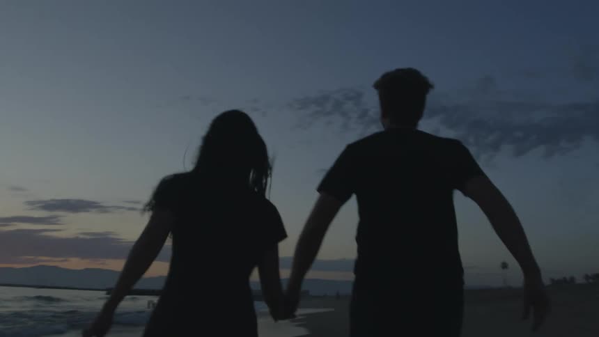 A young man and woman holding hands as they run toward the ocean at sunset