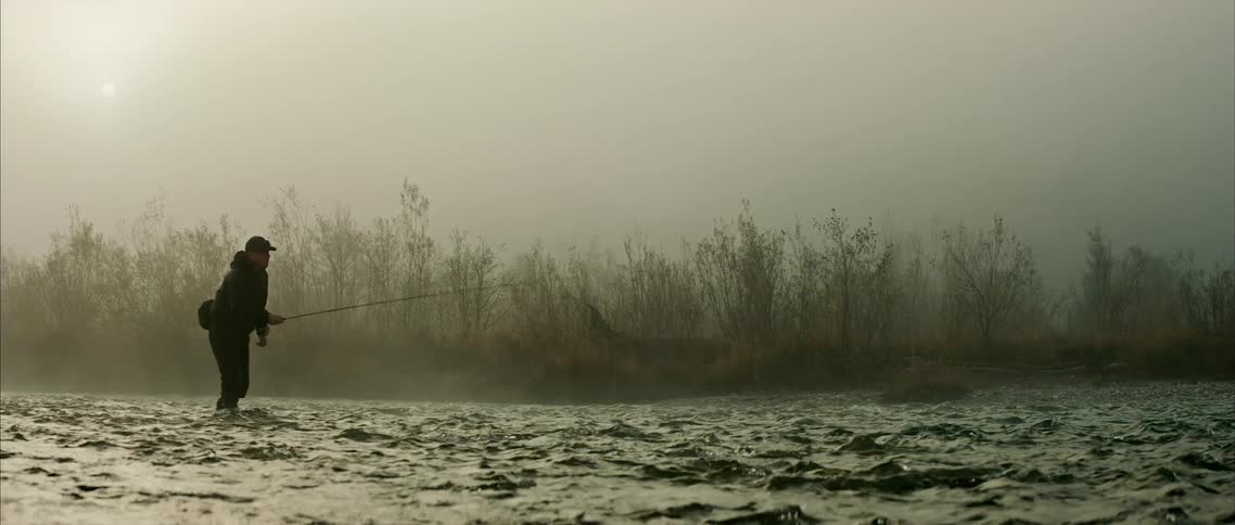 A fisherman casts a fishing line while standing in a river on a foggy morning
