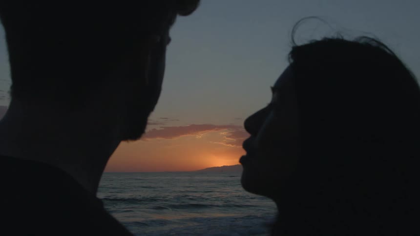 Man with beard and black sweater kisses womans forehead with black straight hair on the coast at sunset