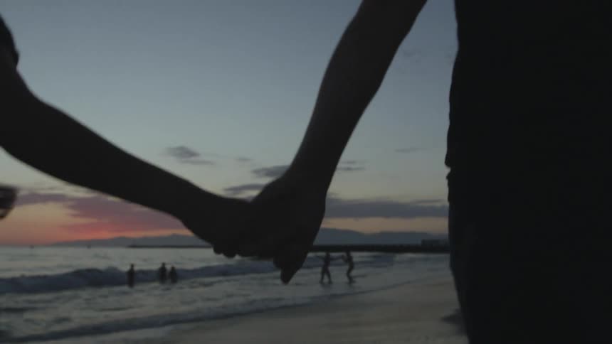 Man and woman walking on the beach next to ocean are silhouetted against the sky at dusk
