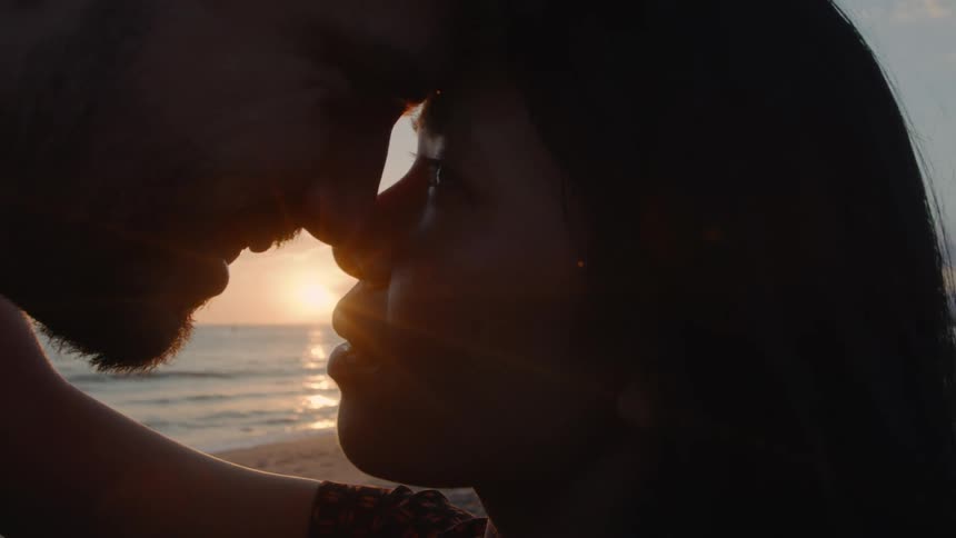Couple stands face to face on the beach as sun sits over water just above the horizon in a red sky