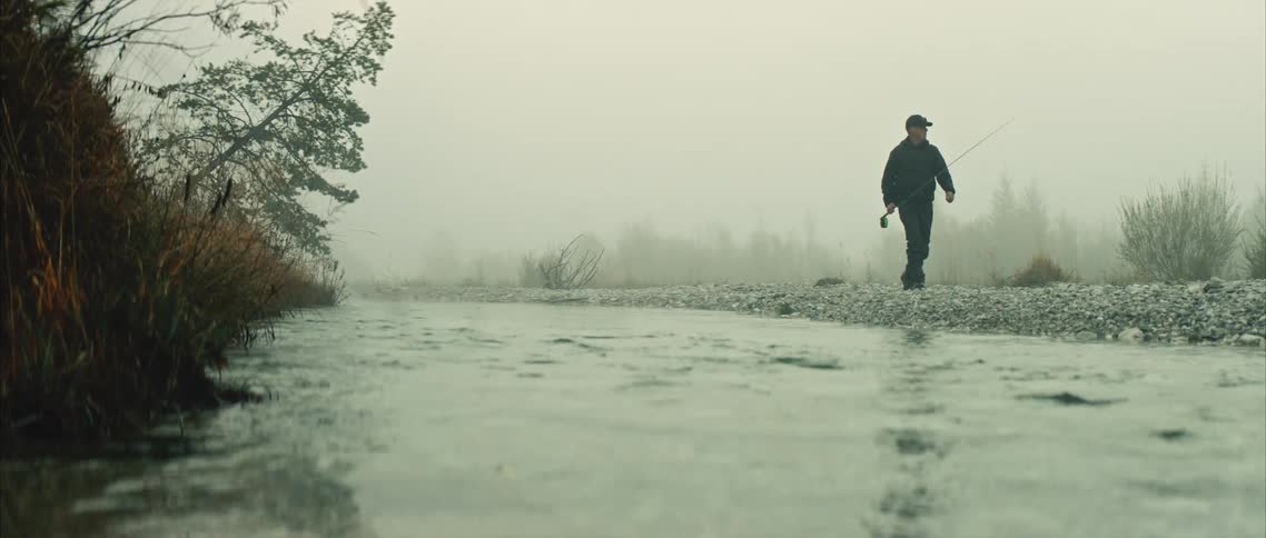 Man with black cap coat and pants walks along river bank carrying fishing rod on cloudy and foggy day