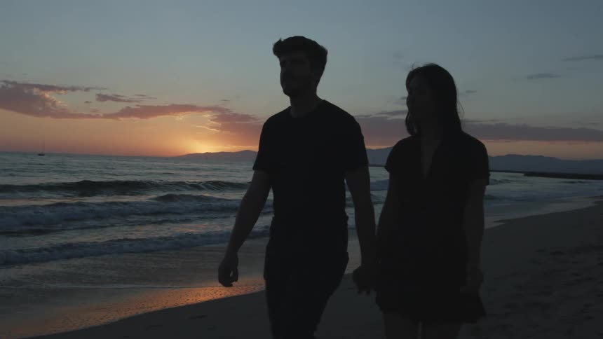 A man and woman walk down the beach holding hands as the sun sets