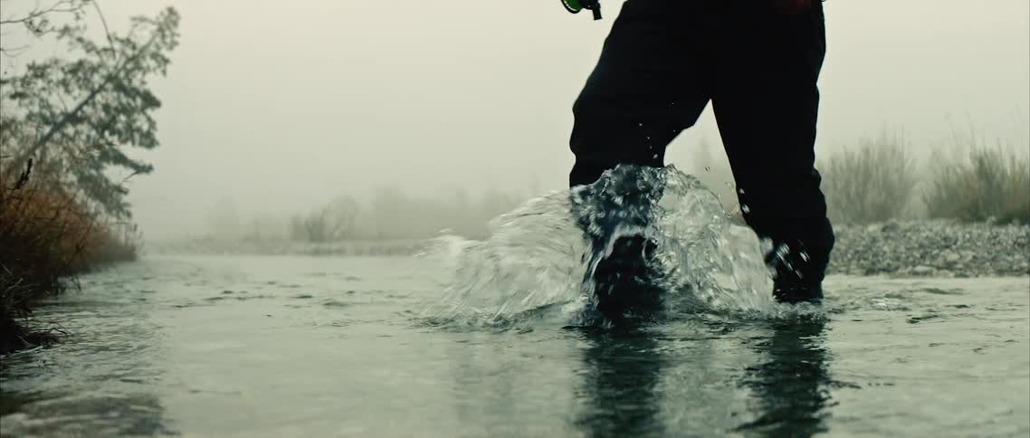 A man is holding a fishing pole and walking through a creek in the wilderness