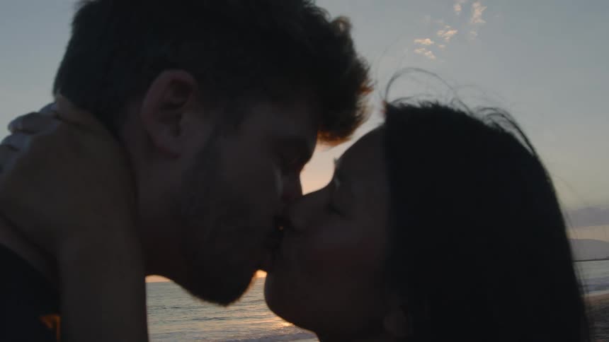 A young couple is at the beach and are kissing by the ocean