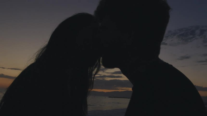 A man and a woman are standing together on a beach and they kiss as the sun sets on the horizon