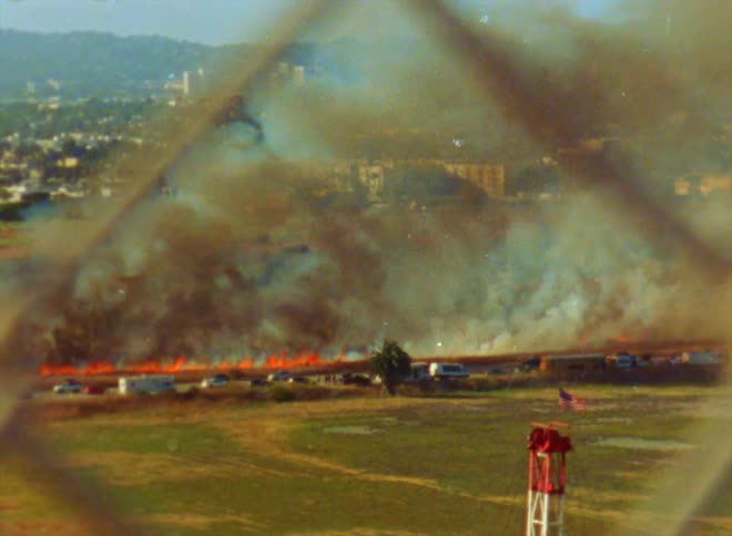 View through a chain link fence of a wildfire burning along the side of the road as traffic moves by and a mountainside neighborhood is in the distance