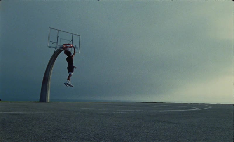 A teenage boy throws a basketball towards the hoop and it bounces then he leaps in the air and slam dunks it through the hoop