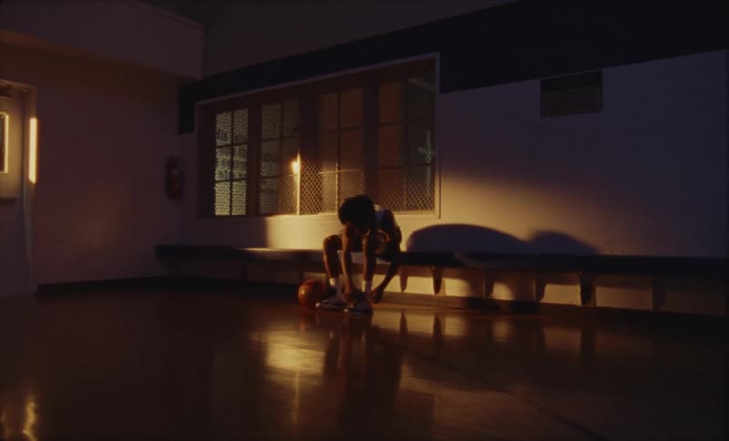 Young man ties shoes while sitting on a bench inside a dimly lit basketball court