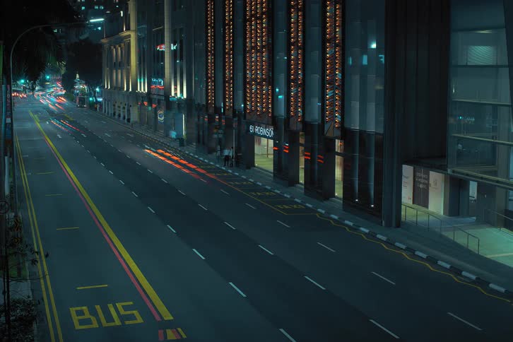 Lights on vehicles shine as they travel on a road between dimly lit downtown buildings as people walk on the sidewalk