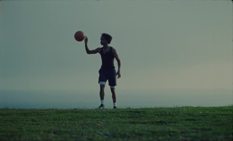 Basketball player in black undershirt and purple shorts does tricks with basketballon grass