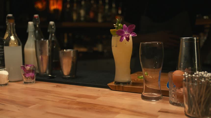 Barman wearing black shirt and apron puts cocktail at the bar next to alcohol bottles