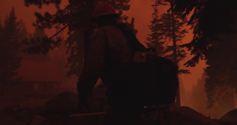 A firefighter walks through a smoky forest with fires burning towards a house