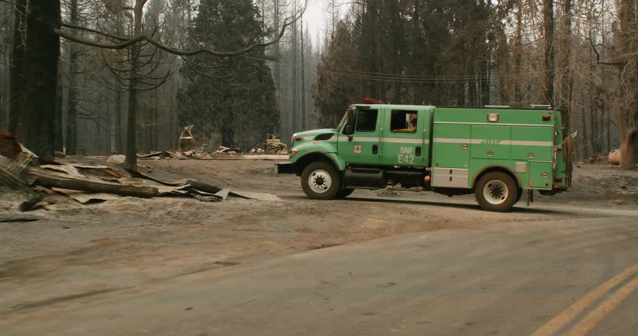Green firefighter truck parked on a rural road near a burned house in the forest