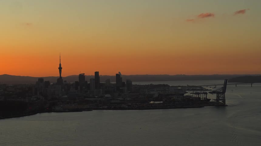 Silhouette a downtown skyline with a tower and spire among the buildings along the coast under an orange sunset sky and mountains in the distance