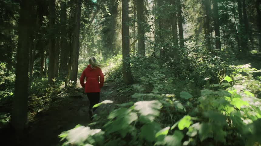A woman is running on a trail in the woods as the sun shines brightly through the trees