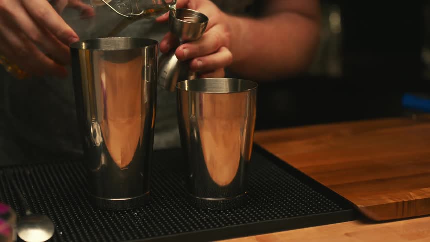 Two metal mixing cups sit on the bar as the bartender adds a shot