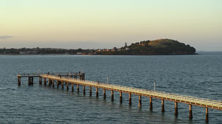 People move along pier extending into water across from town under a red tinted sky