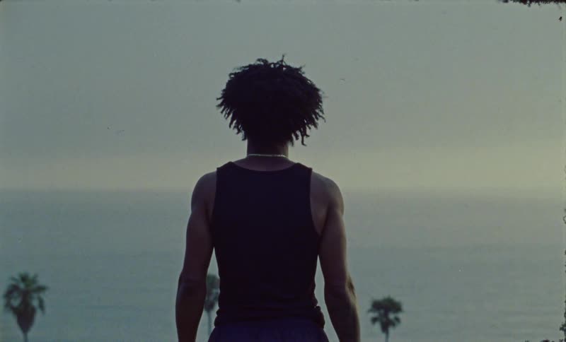 Man with curly hair and black undershirt watches ocean on cliff overlooking ocean