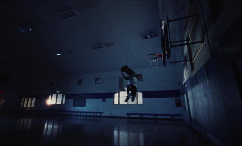 The silhouette of a young man throwing up a basketball and dunking it in a school gym at night