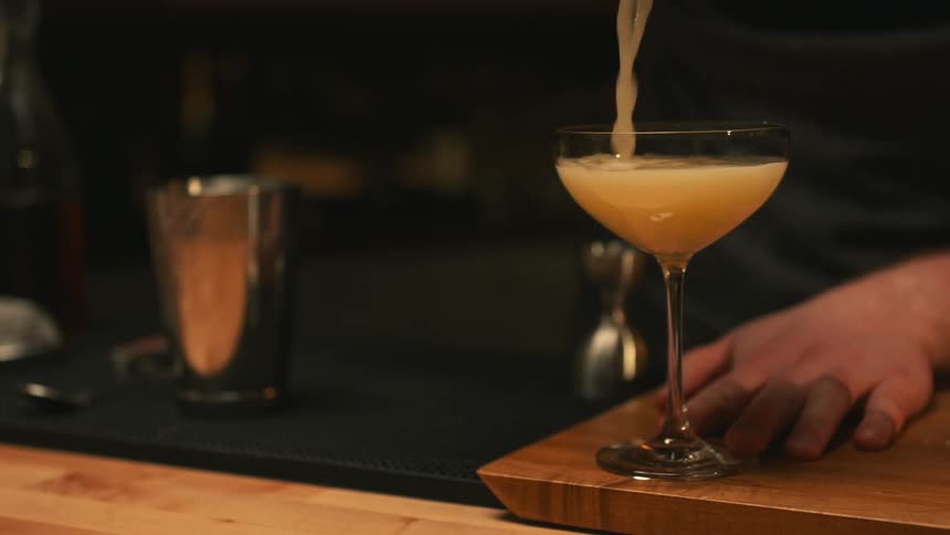 A bartender pours a mixed drink from the metal mixing cup into the glass
