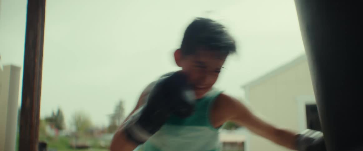 A boy wearing boxing gloves is outside on his porch practicing boxing hitting a punching bag