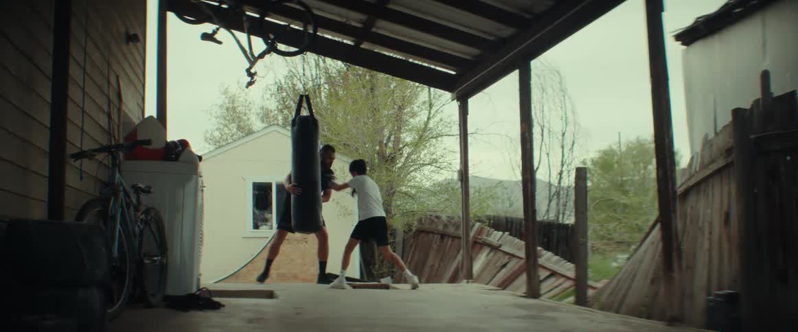 A boy is outside on his porch wearing boxing gloves and hitting a punching bag as his father holds it and coaches him