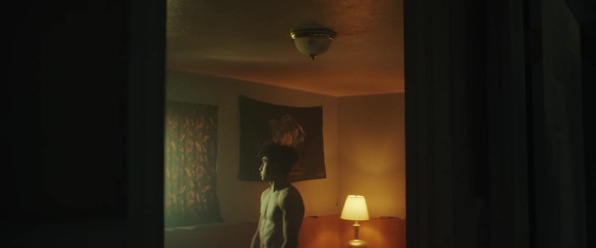 A young man standing in a room looking out a window through the gap between the curtains