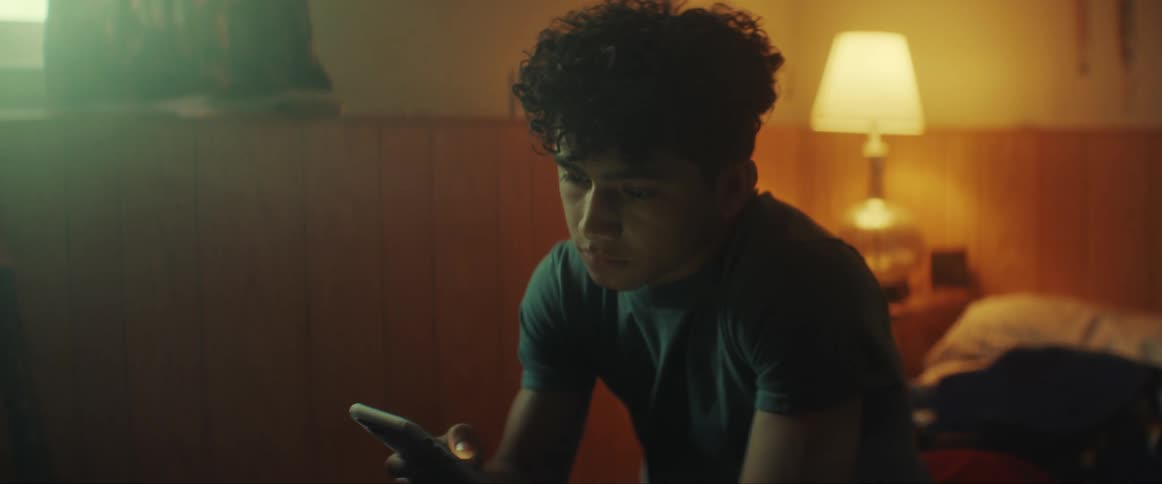 Young teenage boy checks his phone while sitting on the bed in his bedroom