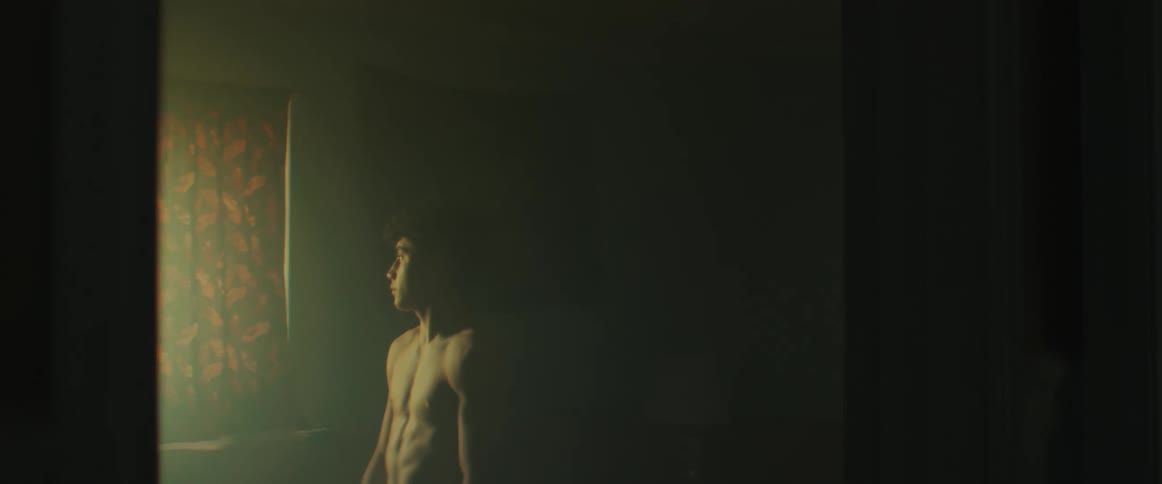 A young man standing in a dark room looking out a window through a gap in the curtains