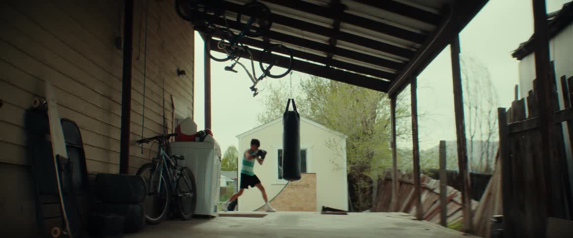 Young sweaty boy practices boxing with a punching bag hanging on the porch in the backyard