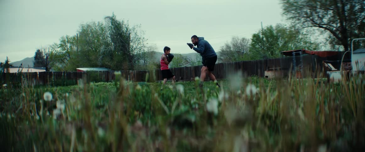 A dad wearing pads trains his son on boxing and corrects his form in the backyard
